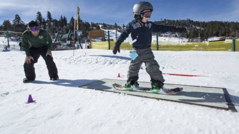 A young snowboarder slides across a rail during a lesson. His instructor looks on.