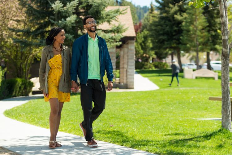 A photo of a couple walking along a paved path at a park with lush green grass and trees in Big Bear Lake