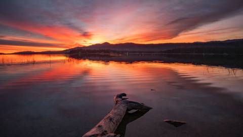 A photo of a bright red-ish pink and orange sunset peering from over the tall mountains, the lake reflecting it like a mirror