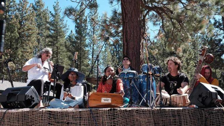 A photo of a band at Big Bear Yoga Festival, some of the band are sitting cross-cross -the lush green forest visible behind.