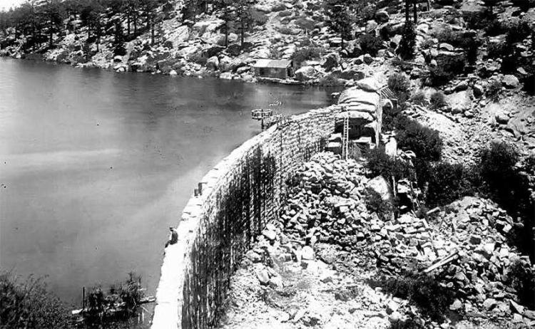 a man sits on the edge of the first, stone Big Bear Lake dam, facing Big Bear Lake prior to 1911